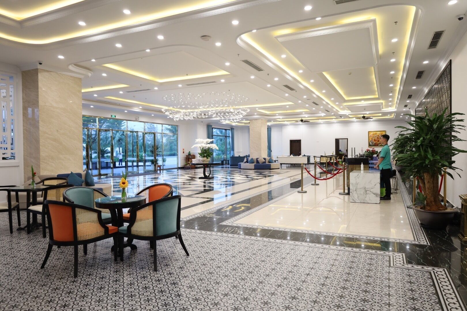 A spacious lobby with a marble floor, a seating area with chairs and tables, a reception desk, and a large potted plant.