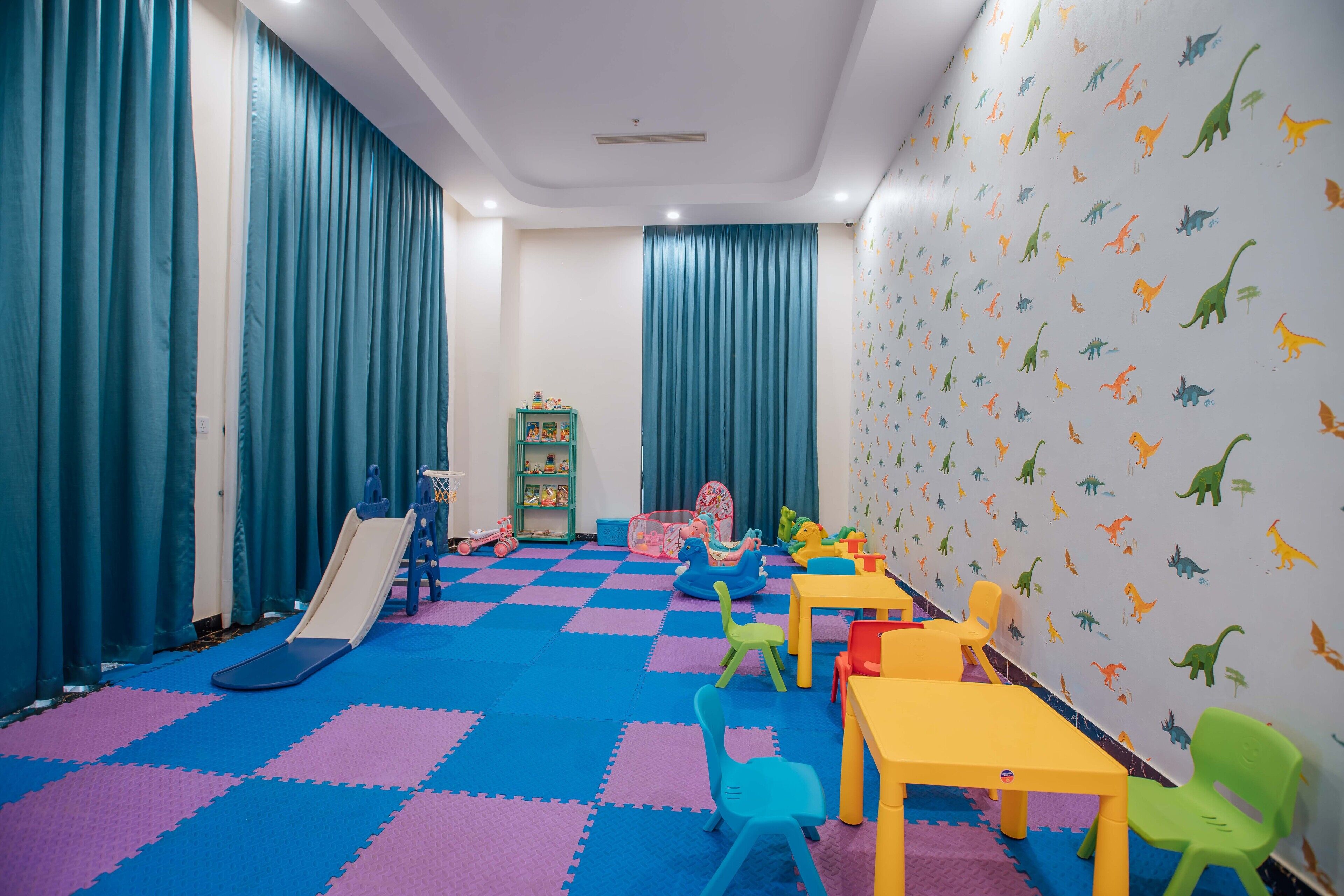 Un cuarto de juegos infantil con un tobogán, sillas coloridas, mesas y calcomanías de dinosaurios en la pared.