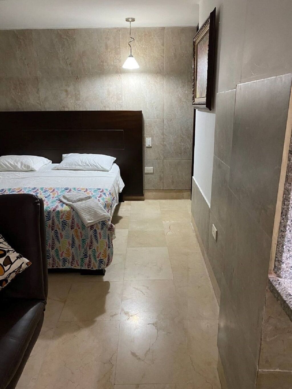 A narrow hotel room with a bed, a small seating area, and a light fixture.