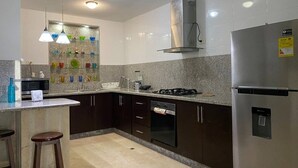 Eine moderne Küche mit dunklen Schränken, einem Edelstahl-Kühlschrank, einem Gasherd und einer Mikrowelle. Drei Pendelleuchten und ein Glastisch mit bunten Gegenständen sind vorhanden.