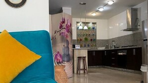 Ein modernes Wohnzimmer mit einem türkisfarbenen Sofa und einem gelben Kissen, eine Küche mit dunklen Schränken und Edelstahlgeräten sowie eine Wand mit floralem Wandbild.