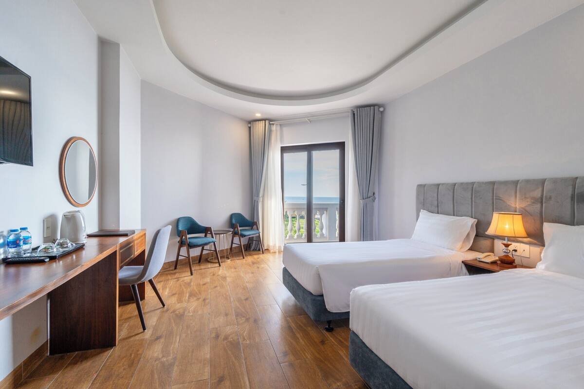 Ett hotellrum med två sängar, ett skrivbord, en stol, ett litet bord och en balkong med gardiner.