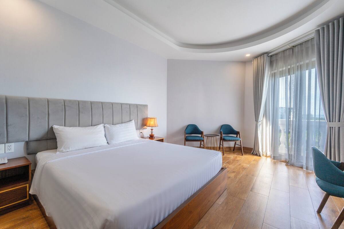 Ett modernt hotellrum med en stor säng, ett nattduksbord, en lampa, två stolar och utsikt över omgivningarna genom tunna gardiner.