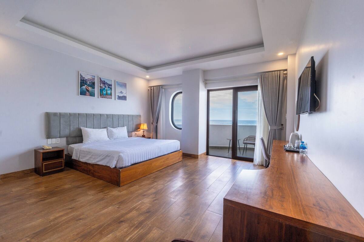 Ett hotellrum med en säng, ett träbord vid sängen, ett skrivbord, en tv som är monterad på väggen och utsikt över havet genom ett fönster.