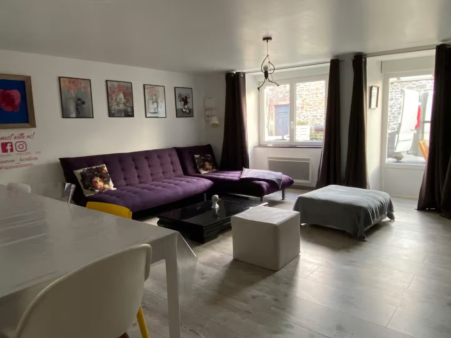 A modern living room with a purple sofa, a white table, and a grey ottoman. There are framed pictures on the wall and a window with curtains.