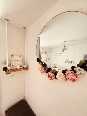Une pièce dotée d’un grand miroir, d’une étagère en bois sur laquelle est posé un panneau encadré, et d’une composition florale fixée au mur.