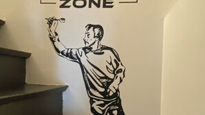 A wall with a sign reading 'NAWENN GAMING ZONE' and a black and white illustration of a person holding a dart.