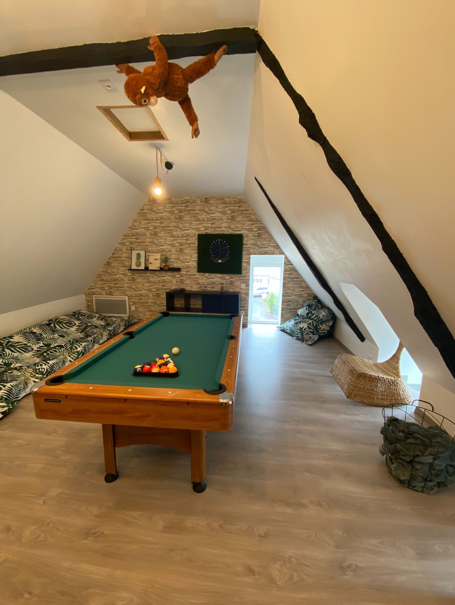 Une pièce avec une table de billard, un jouet en peluche suspendu, un mur en pierre et un plafond en pente.