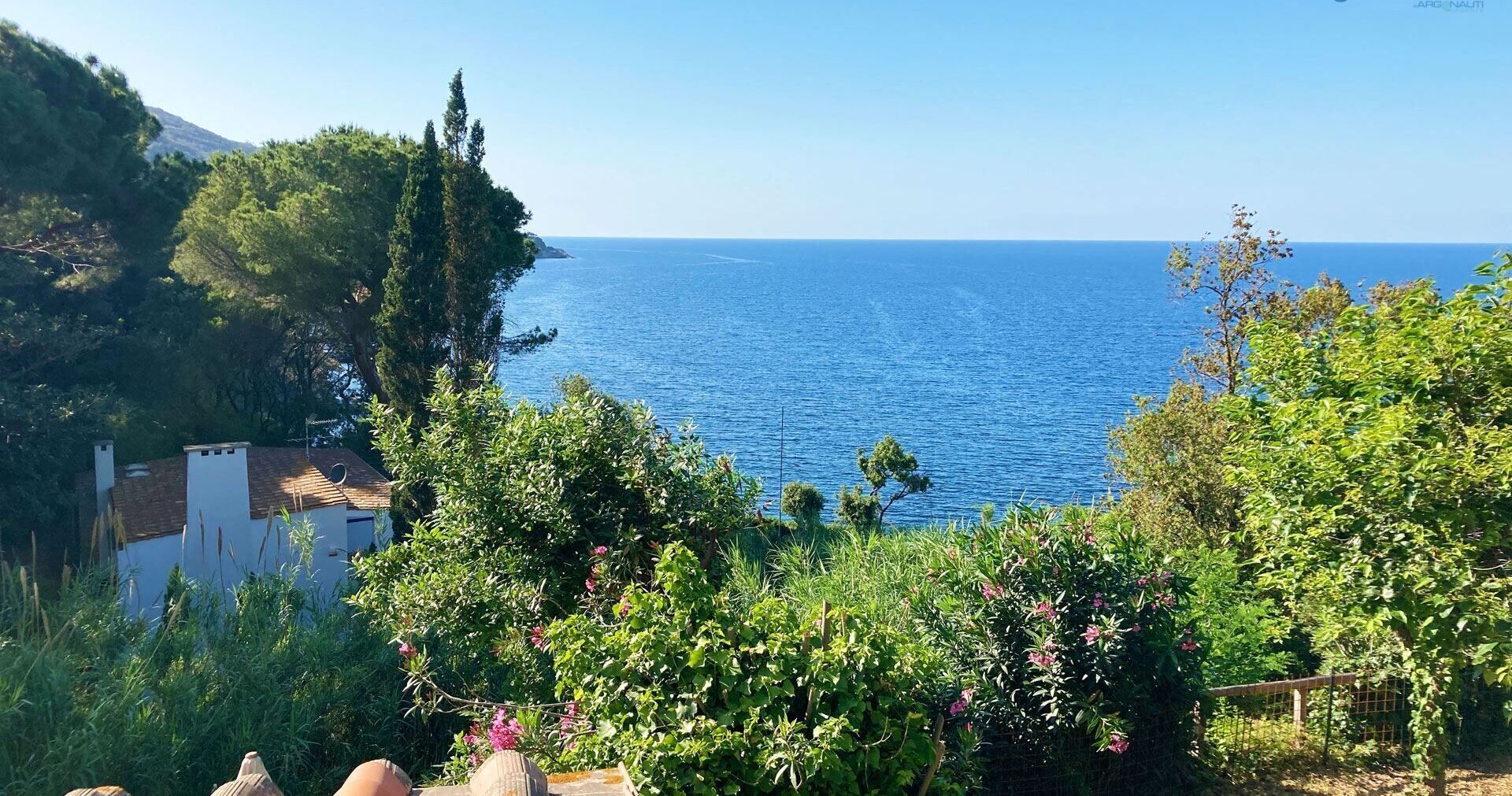 A coastal view with a clear blue sea, greenery, and a white building with a tiled roof.