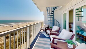 Un balcón con vistas a la playa, amueblado con sillas y una pequeña mesa, y una escalera espiral que conduce a un nivel superior.