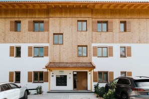 A multi-story building with a wooden facade, a central entrance with a door, and two parked cars.