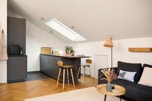 A modern kitchen with a skylight, dark grey cabinets, wooden stools, and a black sofa with a heart pillow.
