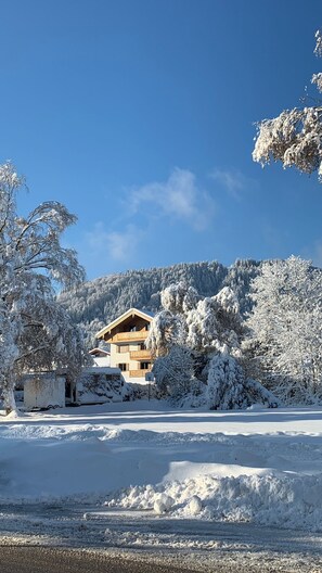 A snow-covered landscape with a wooden house, trees, and a clear blue sky.