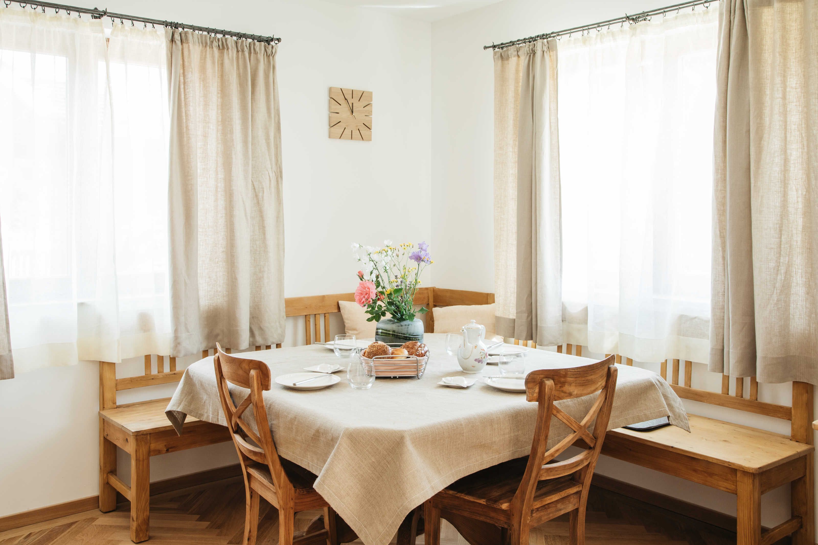 A dining room with a round table set for four, wooden chairs, a vase with flowers, and a clock on the wall.