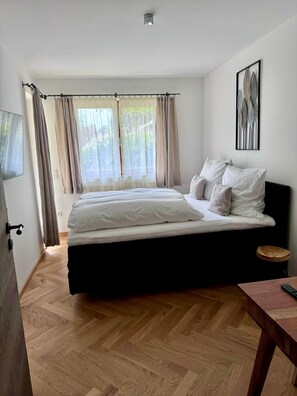 A bedroom with a bed, a bedside table, a window with curtains, a wall-mounted picture, and a wooden floor.