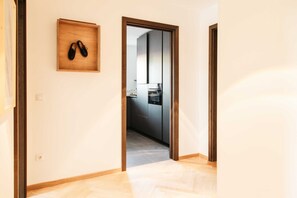 A hallway with a wooden shoe rack, a kitchen visible through an open door, and a herringbone-patterned floor.