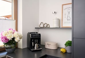 Et køkken med kaffemaskine, en hvid beholder, et indrammet billede og en vase med blomster.