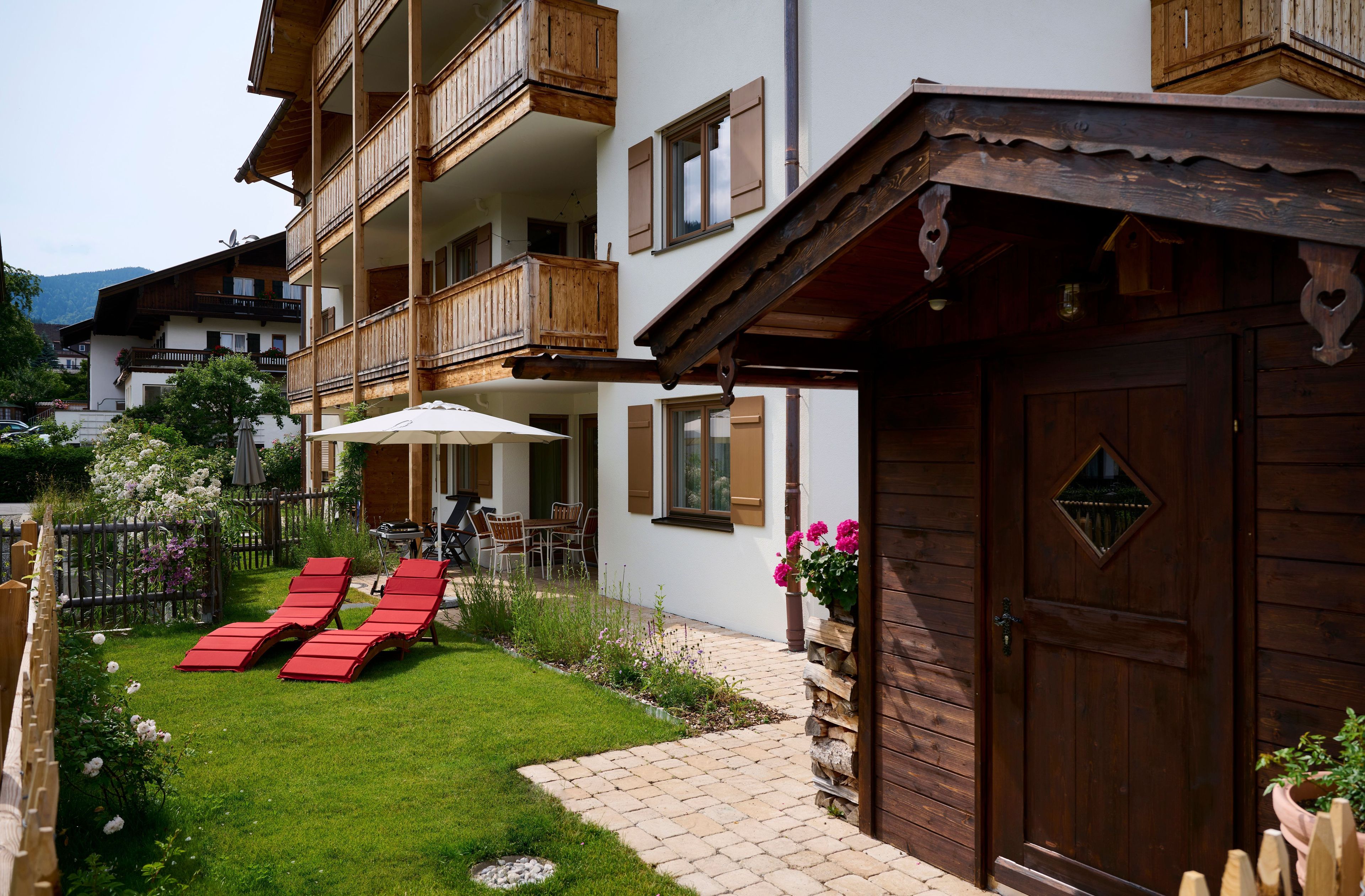 A multi-story building with wooden balconies, a paved patio with red lounge chairs, and a wooden door.