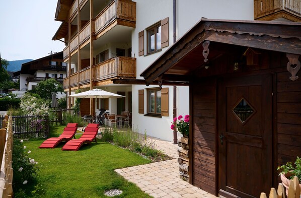 A multi-story building with wooden balconies, a paved patio with red lounge chairs, and a wooden door.