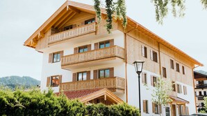 A multi-story building with wooden balconies, a red-tiled roof, and a view of the surrounding landscape. - Ferienappartement Haferlschuh - Alpenseensucht Natur-ferienappartements (Bad Wiessee)