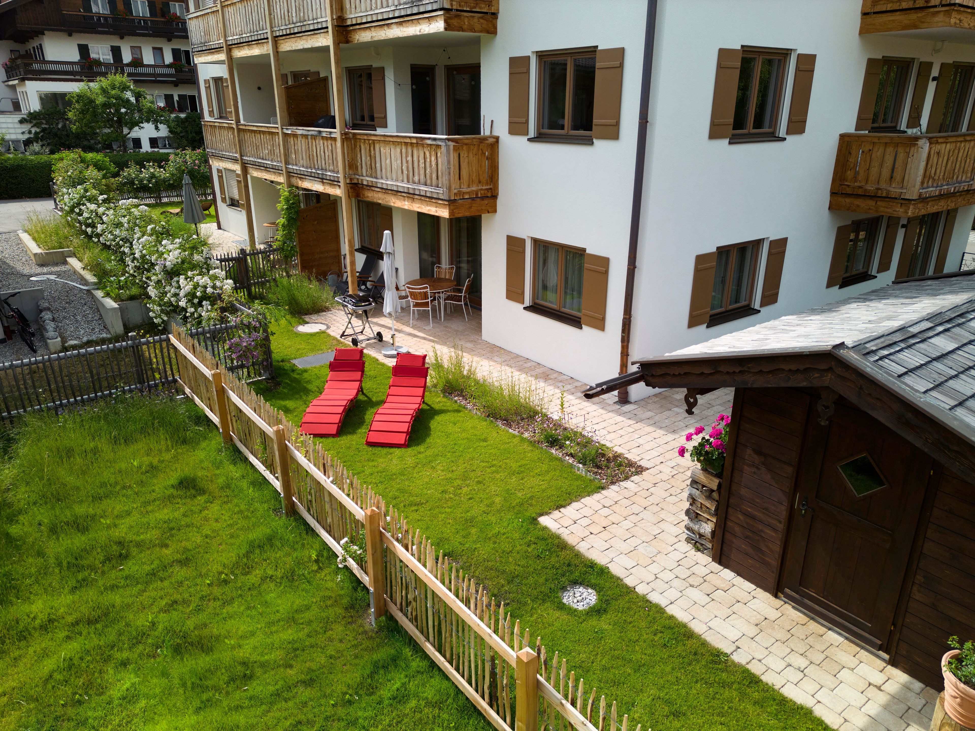 Un immeuble résidentiel doté d’un balcon, d’un petit jardin avec des fauteuils de jardin rouges et d’une terrasse dallée.