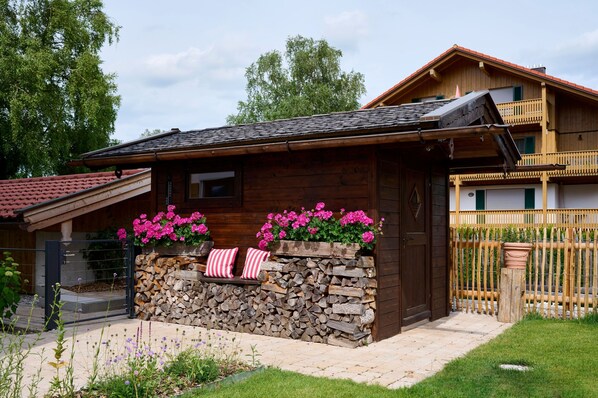 A wooden cabin with a stone wall, a balcony, and a garden with flowers.