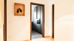 A hallway with a wooden shoe rack, a kitchen visible through an open door, and a herringbone-patterned floor. - Ferienappartement Haferlschuh - Alpenseensucht Natur-ferienappartements (Bad Wiessee)