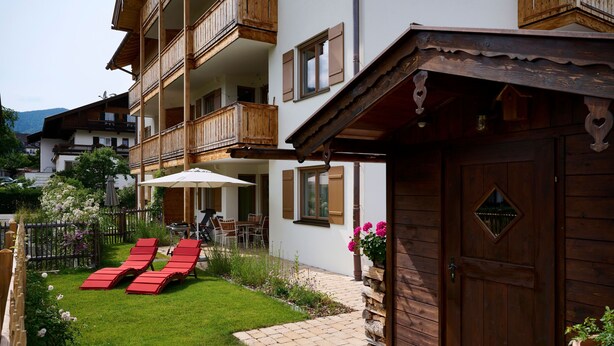 A multi-story building with wooden balconies, a paved patio with red lounge chairs, and a wooden door. - Ferienappartement Haferlschuh - Alpenseensucht Natur-ferienappartements (Bad Wiessee)
