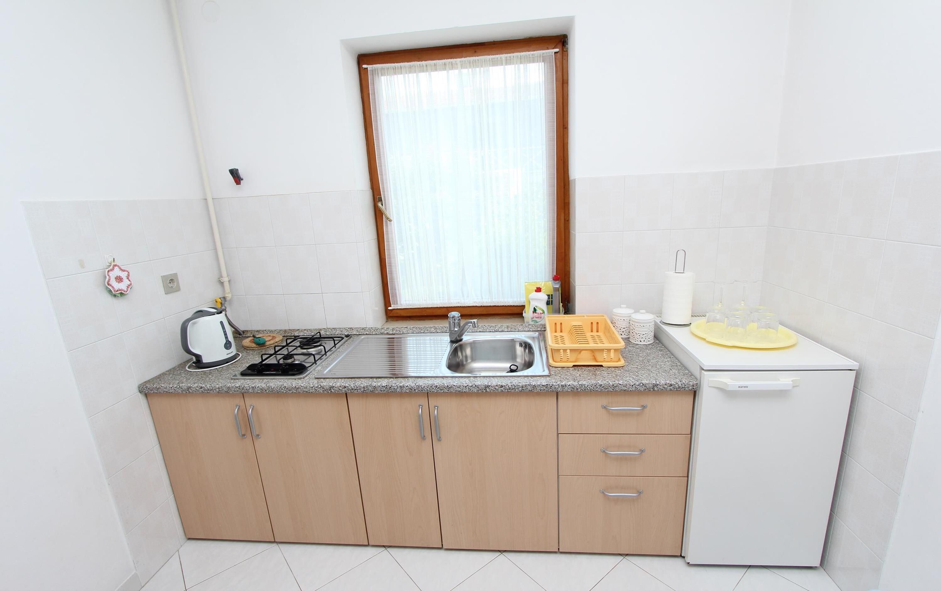 Cozinha compacta com fogão a gás, pia e pequena geladeira. Há uma janela com cortinas leves, uma chaleira e alguns utensílios de cozinha na bancada.