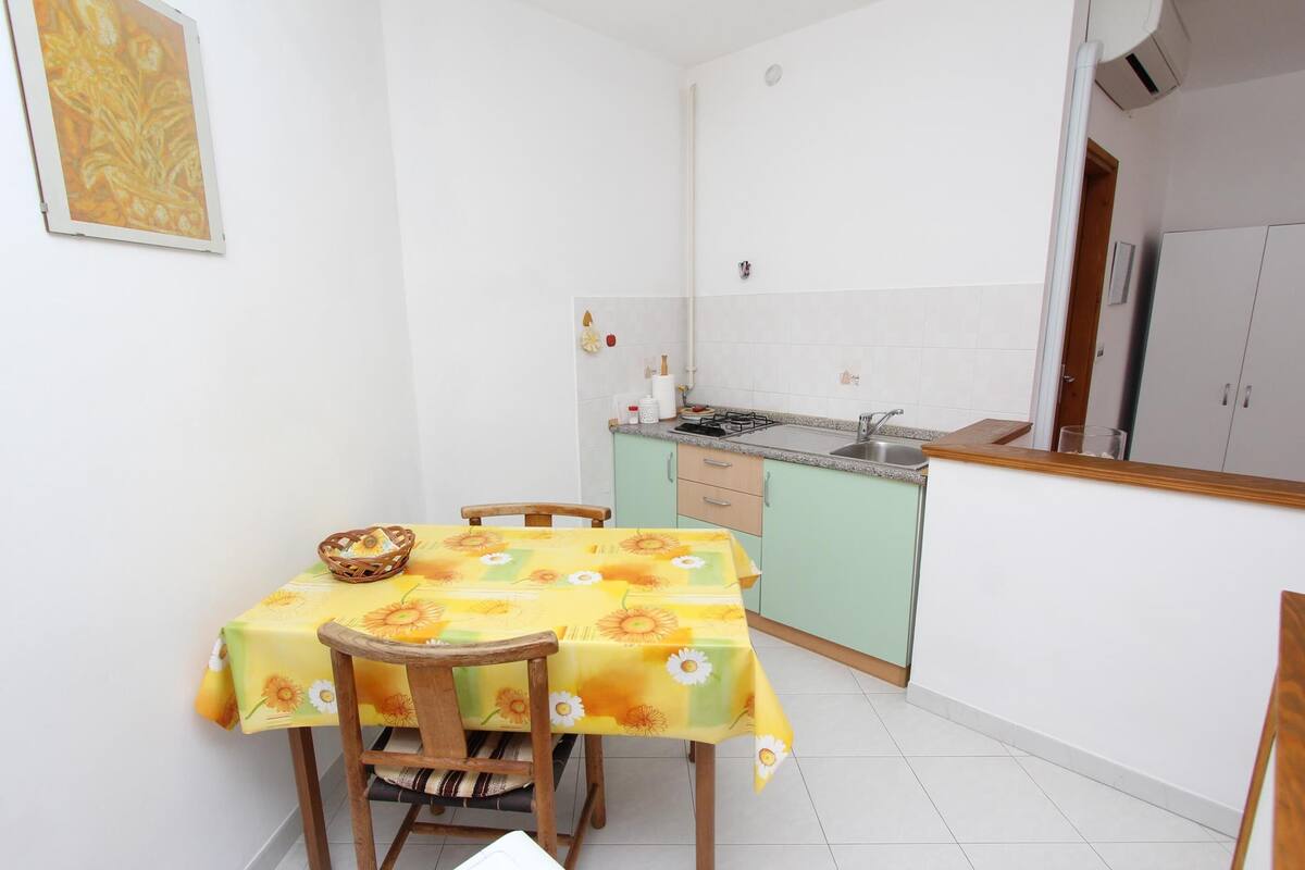 A small kitchen with a dining table covered in a yellow floral tablecloth, wooden chairs, and a basket on the table.