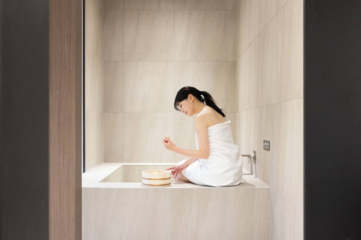 Iemand zit op de rand van een bad in een moderne badkamer met een houten wandpaneel en een klein houten krukje.