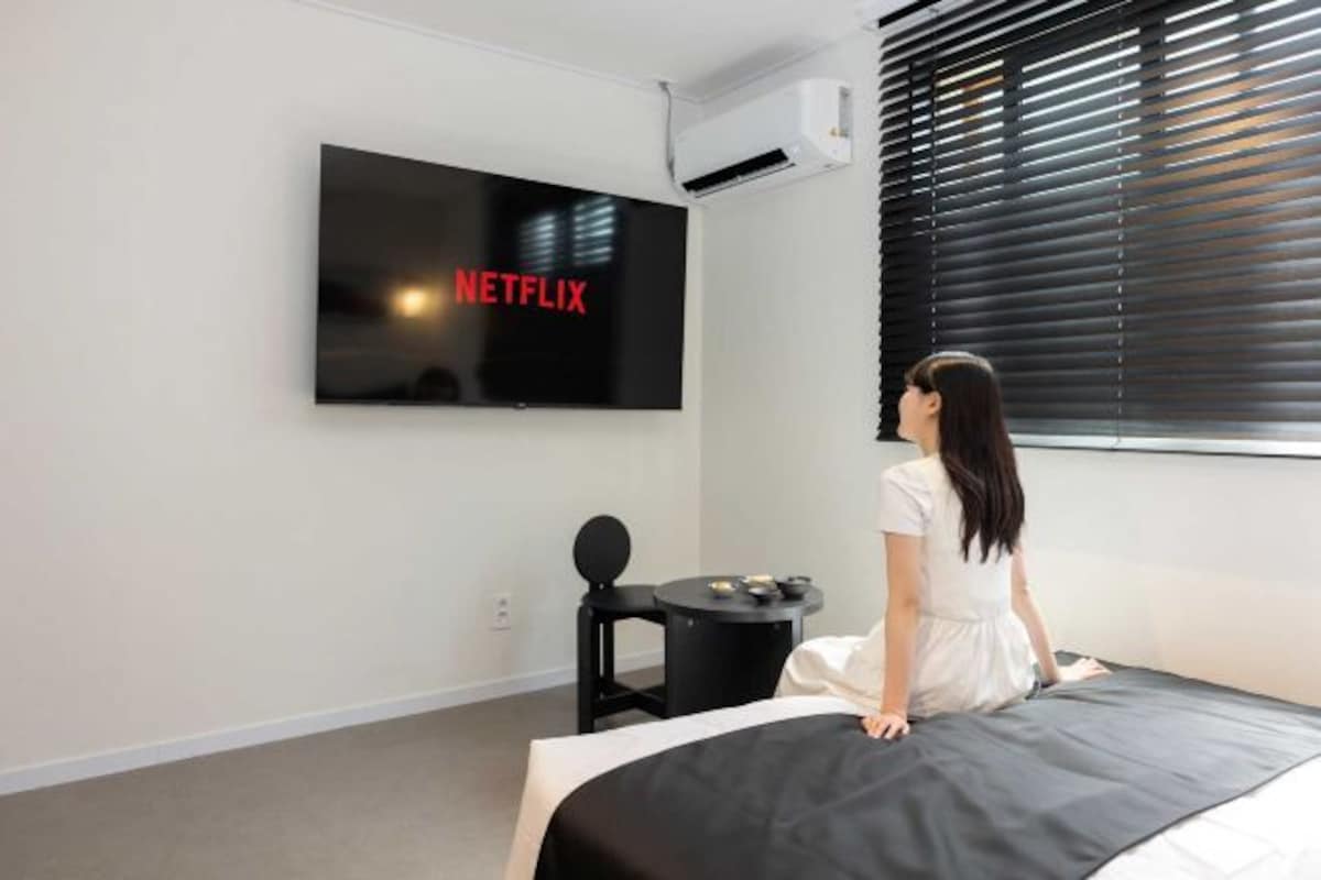 Une chambre d’hôtel moderne dotée d’un grand téléviseur à écran plat fixé au mur, d’une petite table ronde avec une chaise noire et d’un lit avec une tête de lit noire.