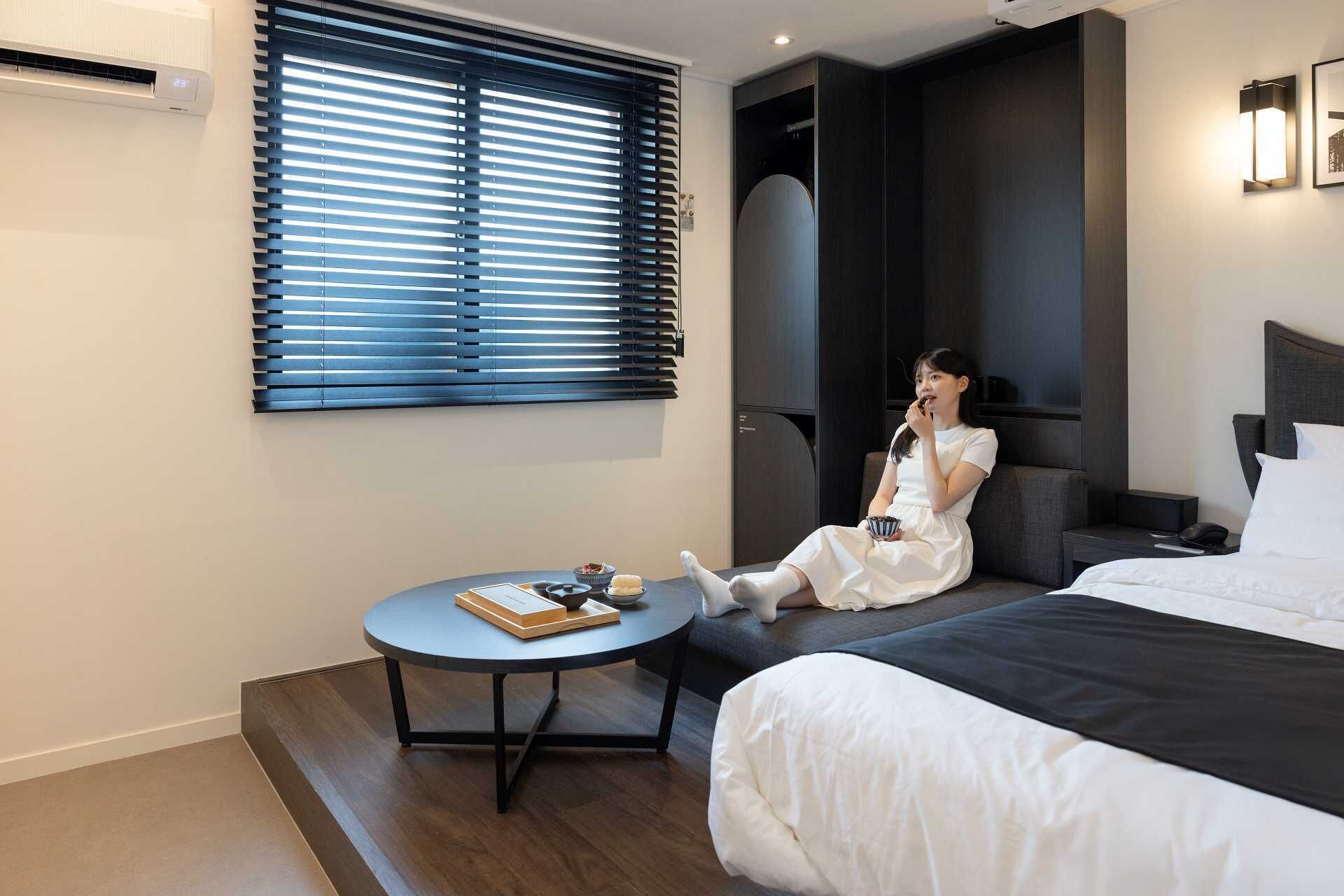 Habitación de hotel con cama, una mesa redonda con un libro y una taza, una ventana con persianas y un aire acondicionado fijado a la pared.