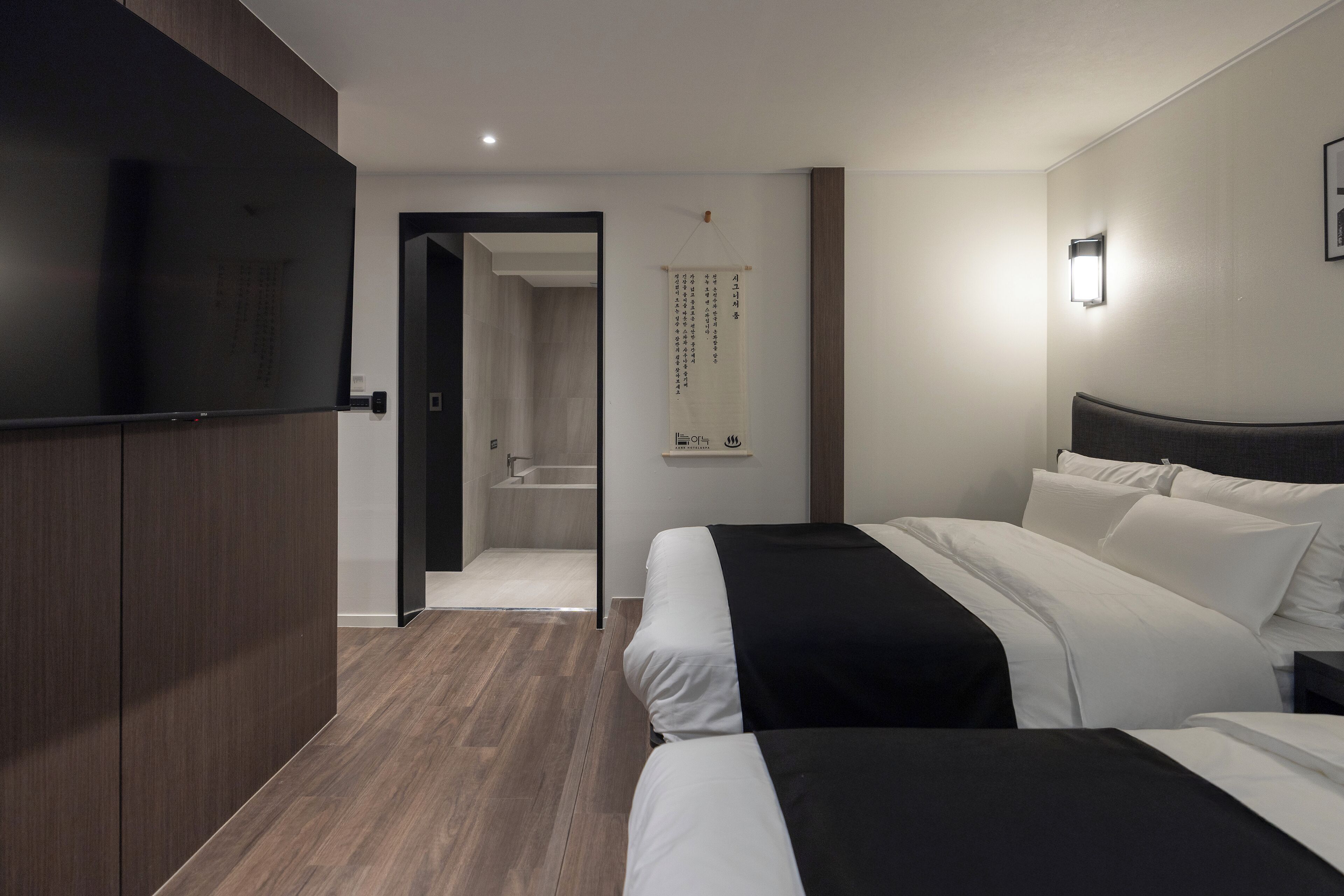 Ein modernes Hotelzimmer mit zwei Betten, einem Flachbildfernseher, einem Holzschrank, einem Badezimmer mit Badewanne sowie einer wandmontierten Lampe.