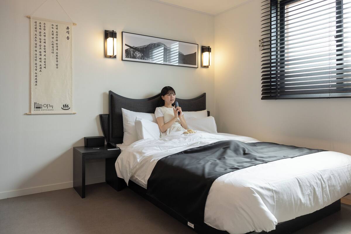 Een netjes opgemaakt bed met zwart-wit beddengoed, een nachtkastje met een lamp, een ingelijst schilderij aan de muur en een raam met zonwering.