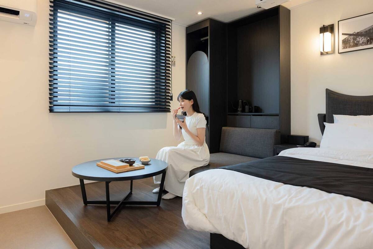 Een moderne hotelkamer met een bed, een klein rond tafeltje met een schaal en boeken, een raam met zonwering, een donkere wand met een lamp en een lijst met een foto.