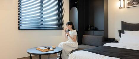 A modern hotel room with a bed, a small round table with a bowl and books, a window with blinds, a dark wall with a light fixture, and a framed picture on the wall.