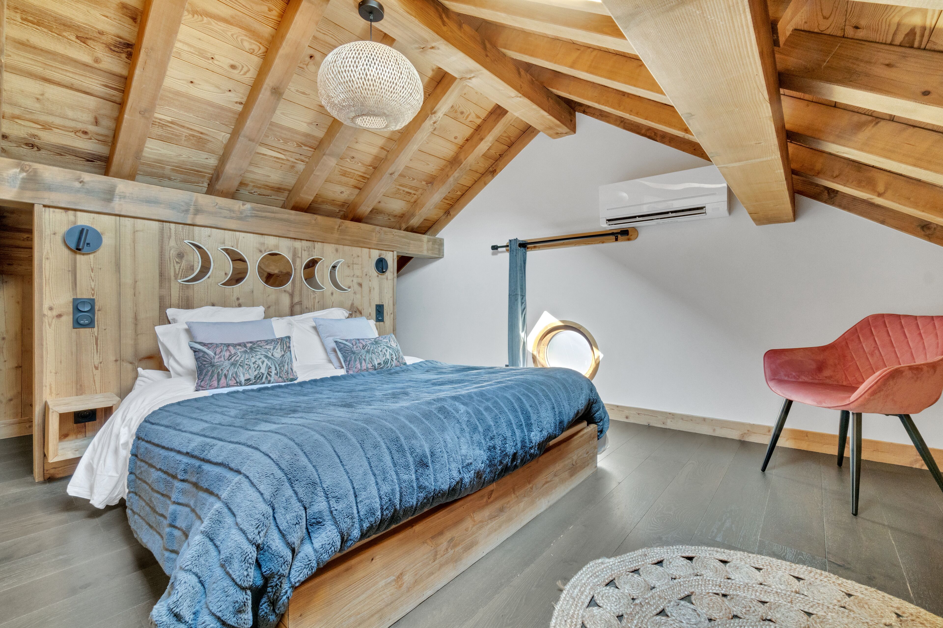 Une chambre avec un plafond en bois, un lit recouvert d’une couverture bleue, une chaise rouge et un tapis tissé.