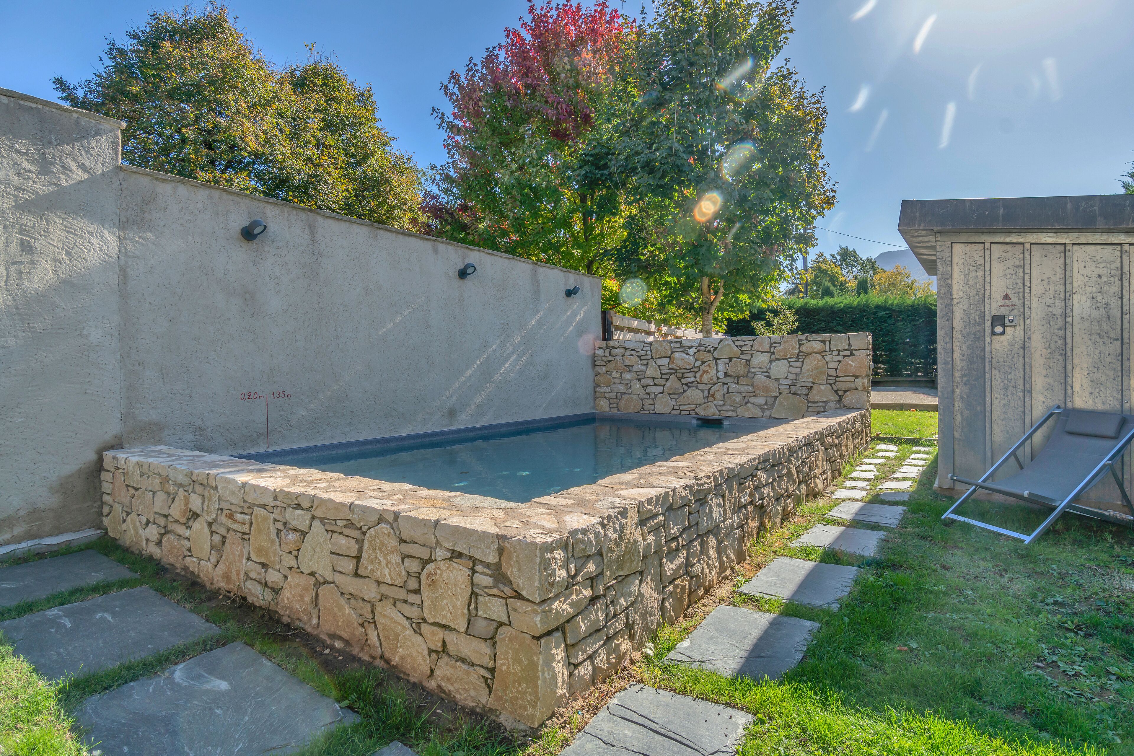 Una piscina con escalones de piedra que la bordean, rodeada por un muro de piedra y una zona con césped.