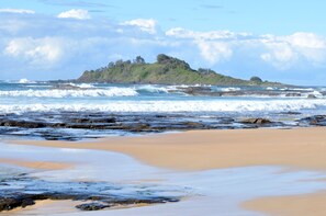 Uma praia com ondas, areal e uma ilha distante coberta de vegetação.