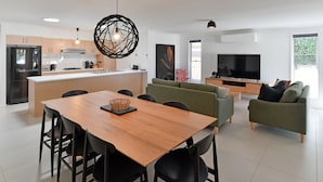 Et moderne køkken med en central ø, et spiseområde med et træbord og sorte stole, en grøn sofa og et fladskærms-TV i en opholdsstue.