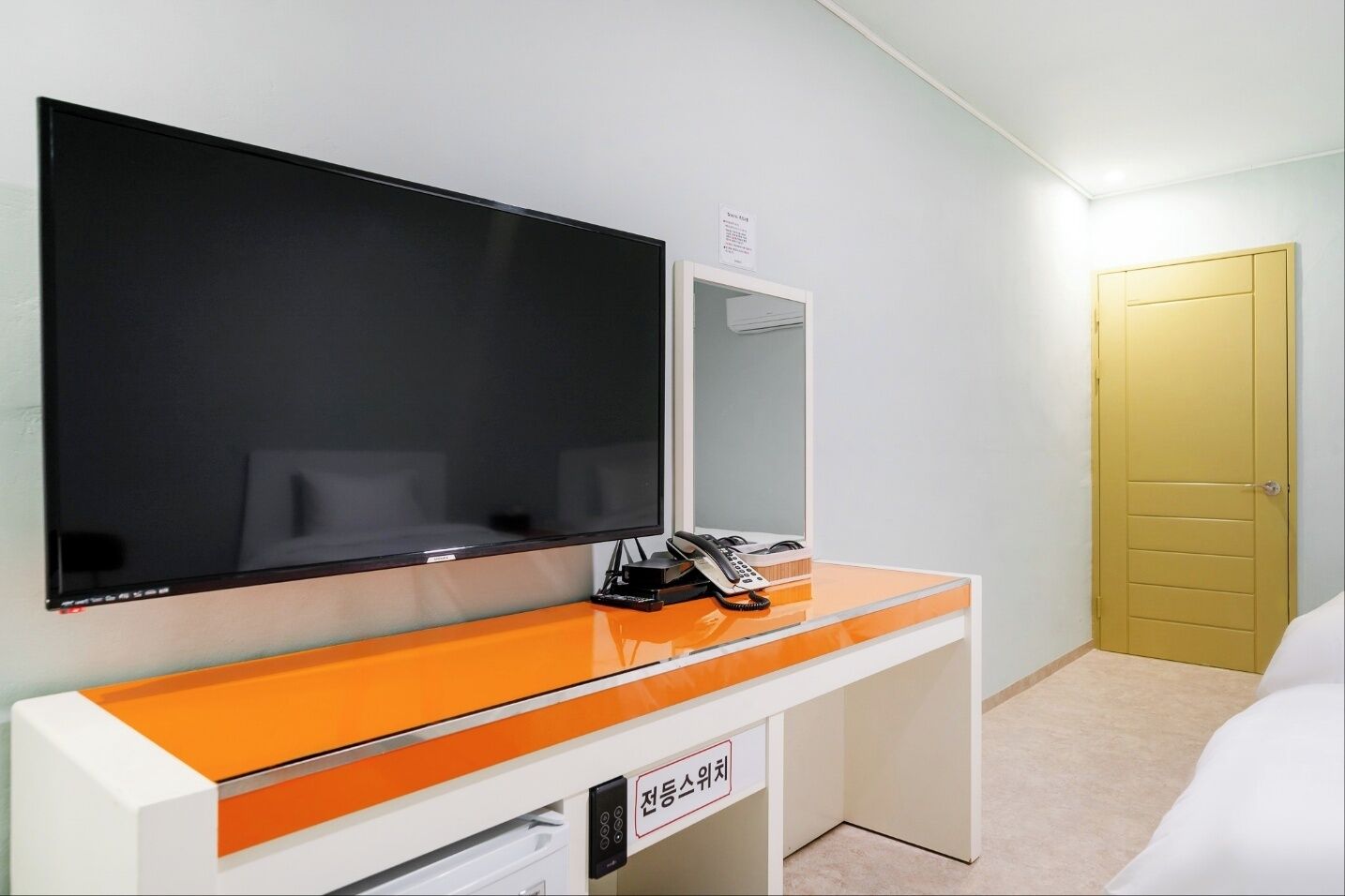 Ein Hotelzimmer mit einem großen Flachbildfernseher, der an einem orangefarbenen und weißen Schreibtisch befestigt ist. Es gibt ein Telefon und einen Spiegel. Im Raum befindet sich eine gelbe Tür und ein weißes Bett.