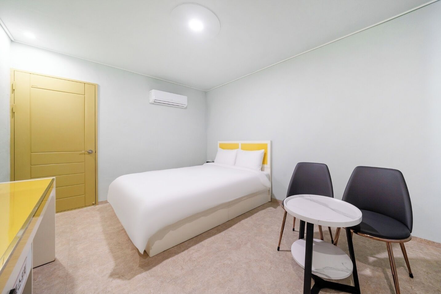 A hotel room with a bed, two chairs, a small table, a yellow door, and an air conditioning unit.