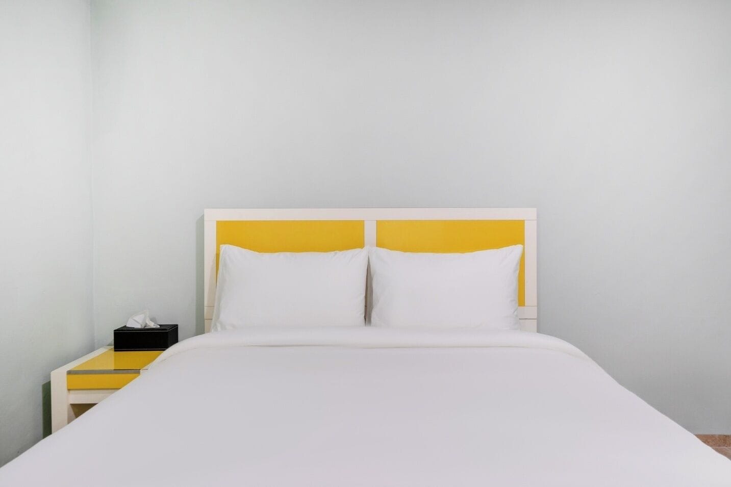 A bed with a white mattress and two pillows, a bedside table with a tissue box, and a yellow headboard.