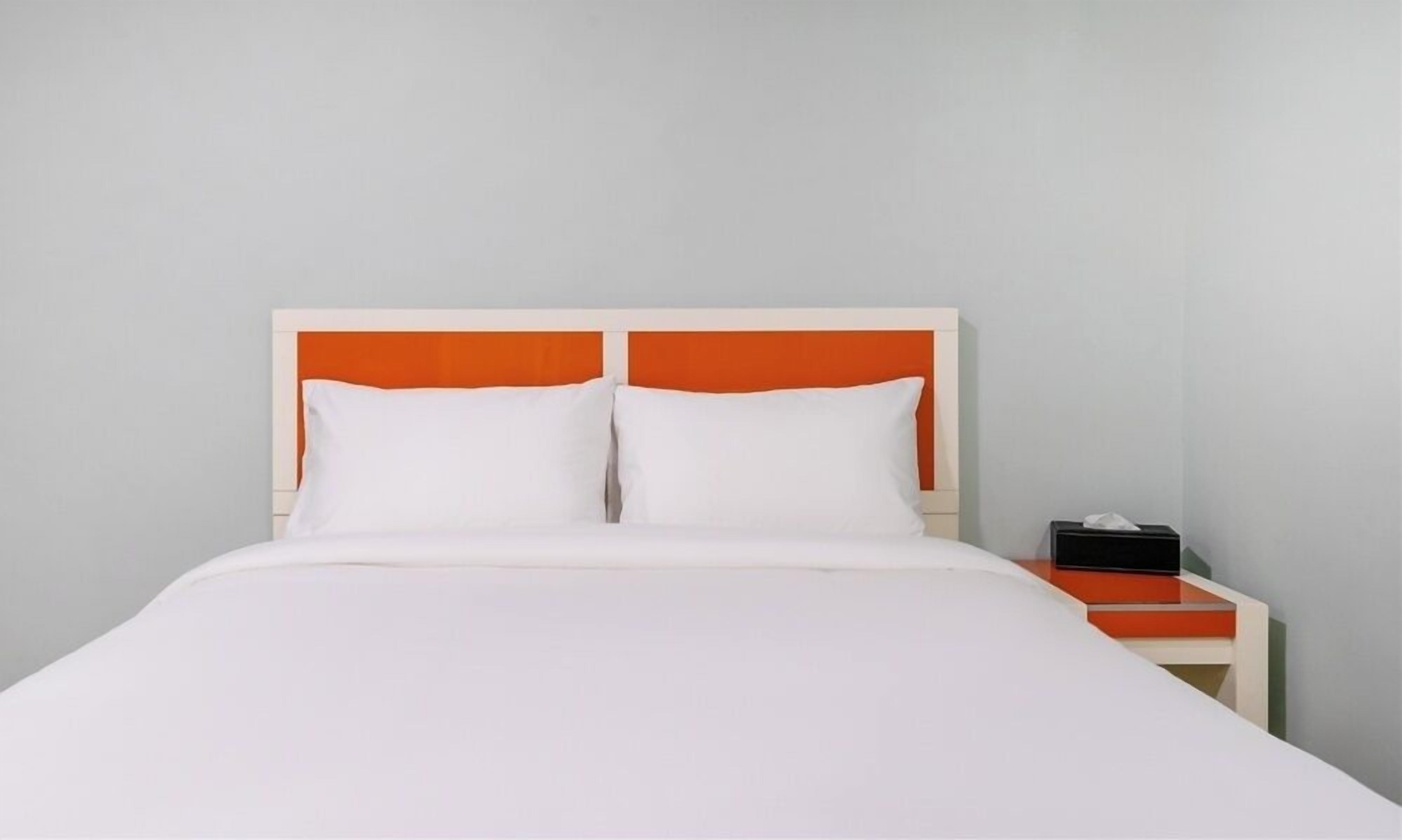 A bed with white bedding and an orange headboard, a bedside table with a black object, and a plain wall.