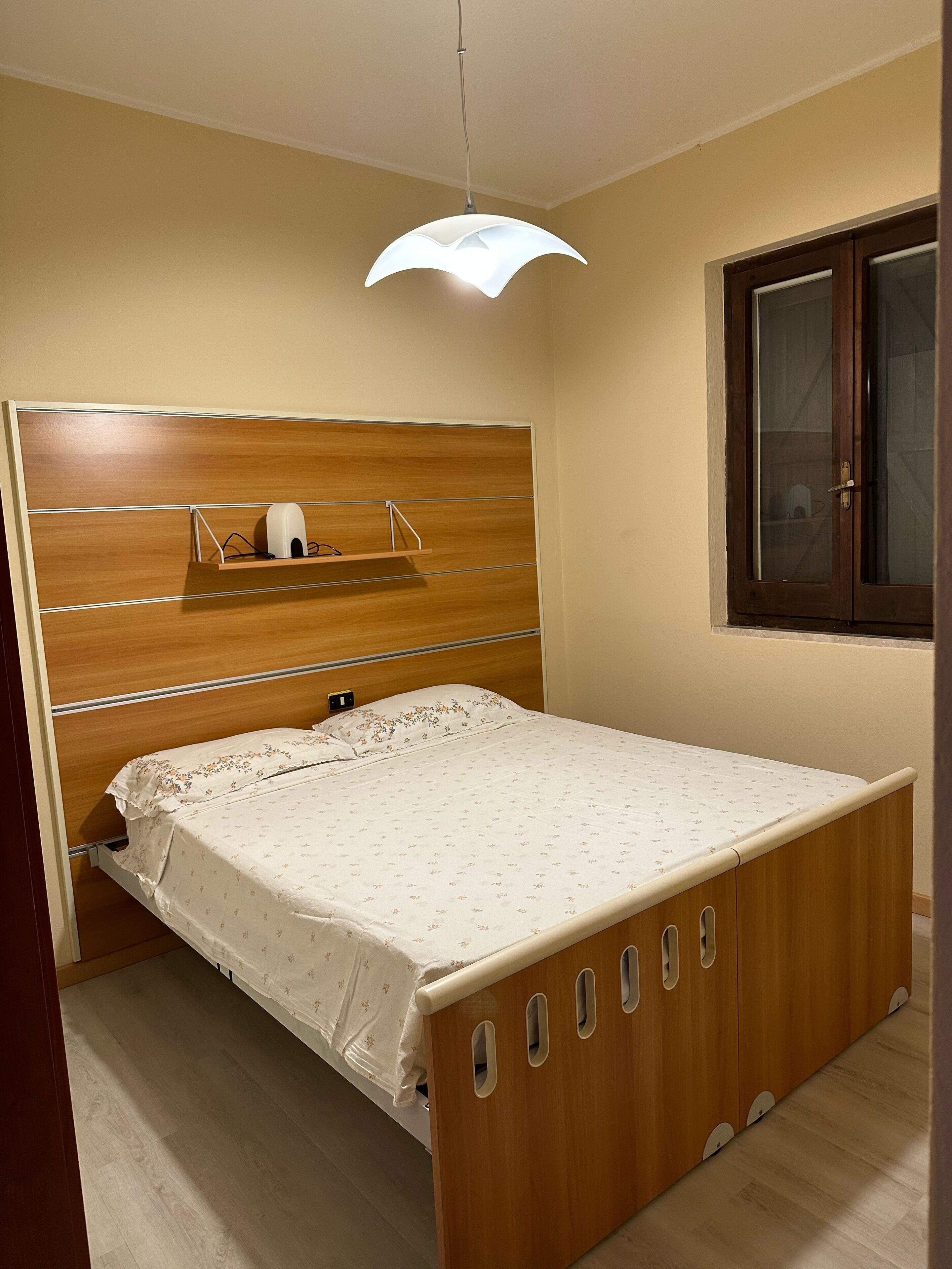 Une chambre à coucher avec un lit en bois, une tête de lit dotée d’étagères, une table de chevet et une fenêtre avec un cadre en bois.