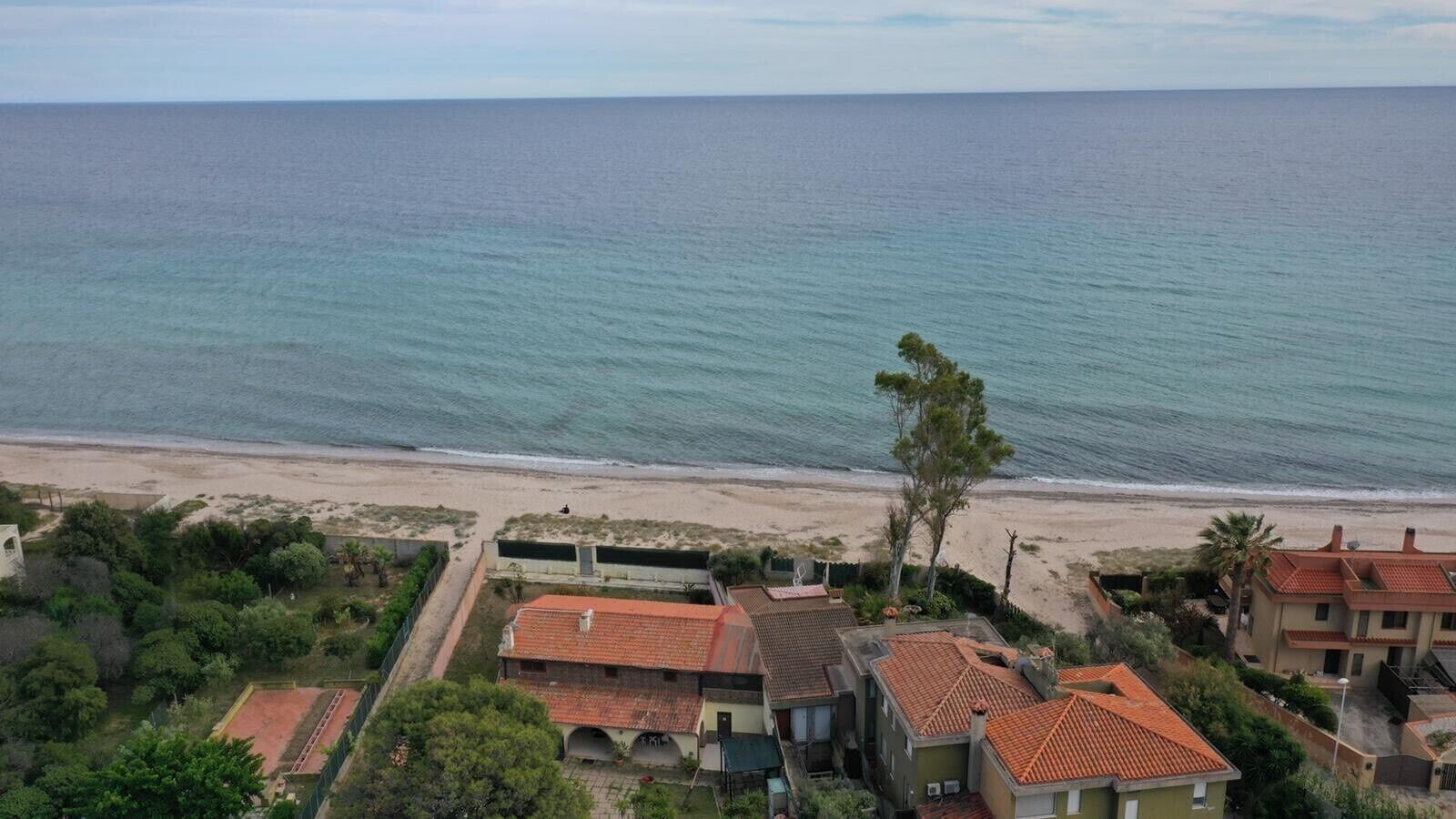 A coastal residential area with houses having red-tiled roofs, a beach in the background, and a clear view of the sea.