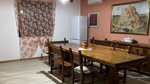 Un comedor con una mesa y sillas de madera, ventilador de techo, cortinas con motivos florales, refrigerador y un cuadro en la pared.