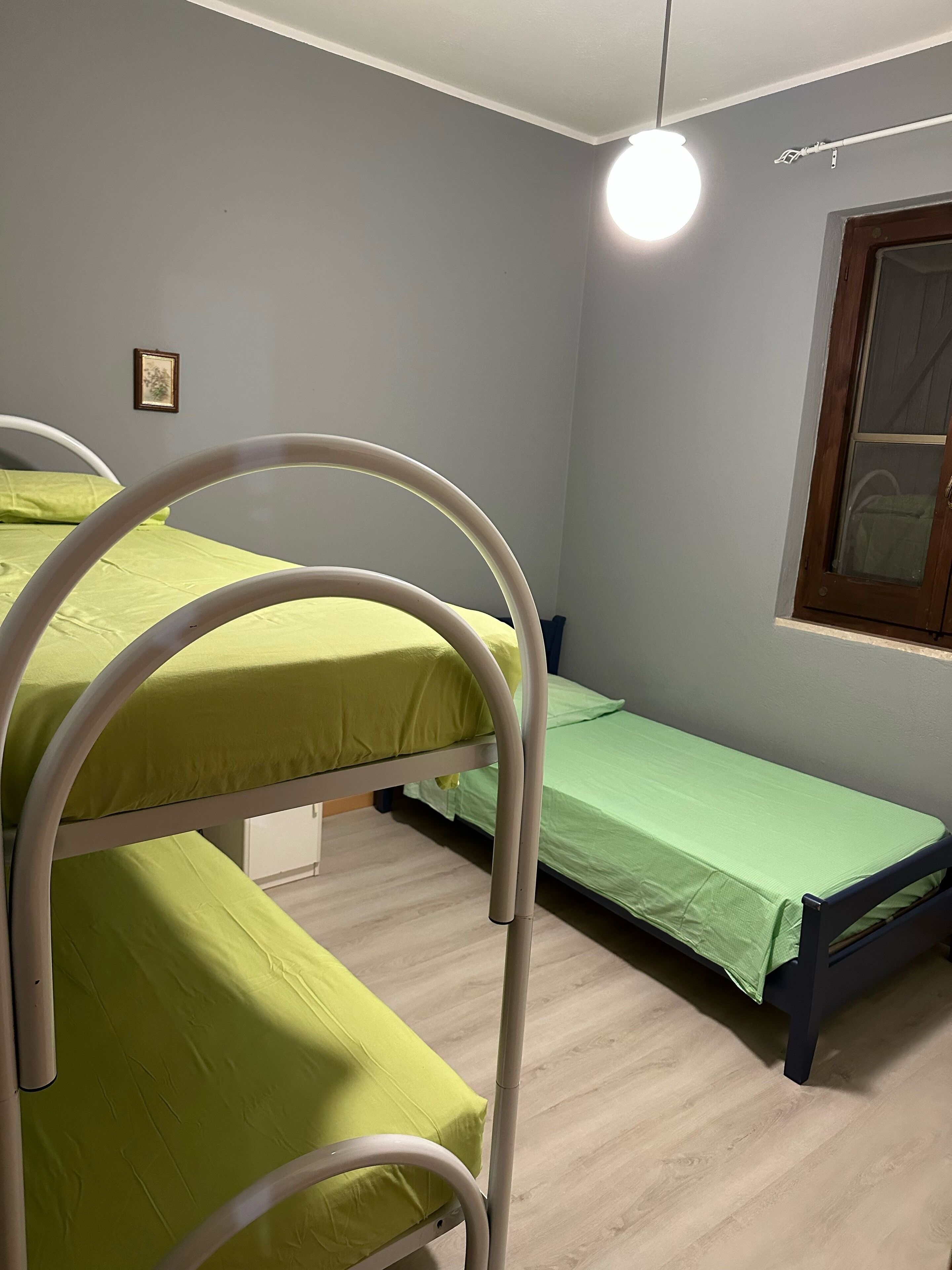Una habitación con literas, una cama individual, una ventana con marco de madera y una lámpara de pared.