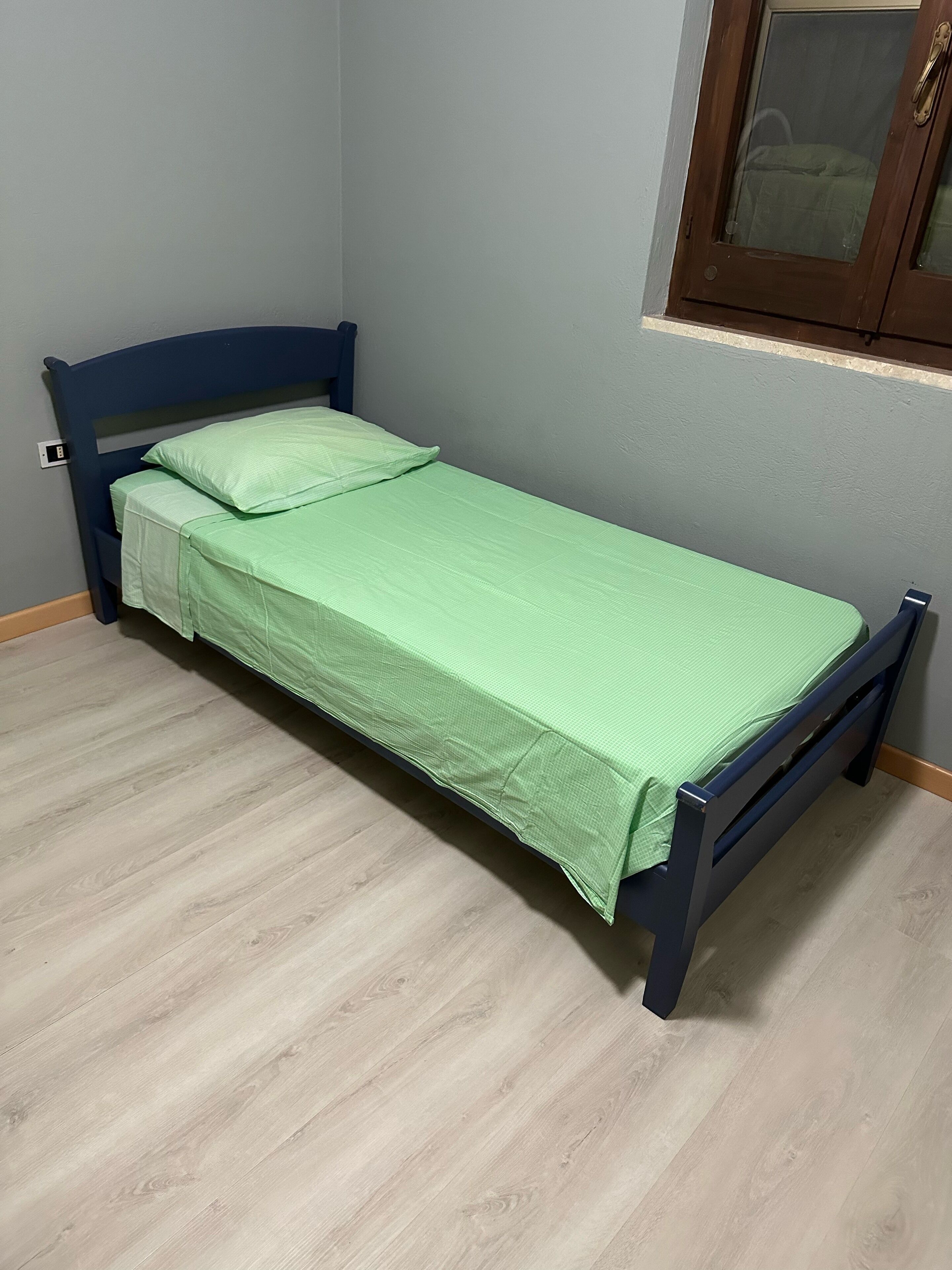 Ein kleines Zimmer mit einem Einzelbett, Holzboden, einem blauen Kopfteil und einer grünen Bettdecke.